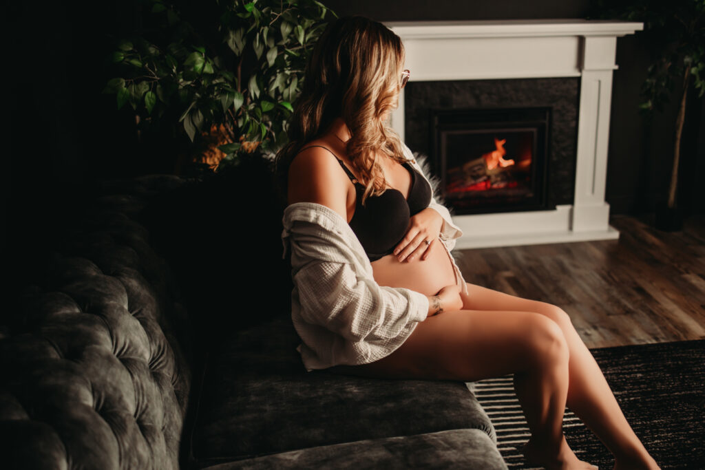 Maternity Boudoir Photography – expecting mother sitting on a couch, gazing at a fireplace.