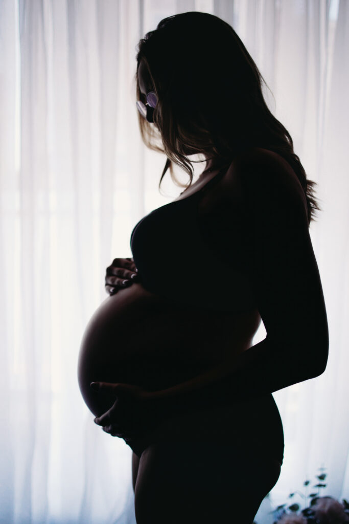 Silhouette of a pregnant woman holding her belly by a soft-lit window.