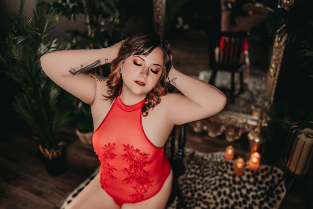 Confident woman in red lace bodysuit during boudoir photoshoot, embracing her femininity with eyes closed and hands in hair, surrounded by candles and elegant decor.